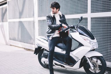 Hızlı sürücü uzakta kendi motosiklet kentsel ortamda otopark süre seyir sevgilisi. Güçlü motoru açık havada beyaz serin skooter üzerinde oturan deri rahat ceket şık genç adam
