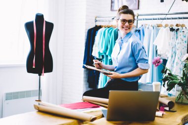 Notlar ölçümlerin showroom koleksiyonu şık giyim konfeksiyon Aksesuar ve netbook masaüstü üzerinde oturan çalışma yaparken kameraya bakarak gözlük gülümseyen kadın Tasarımcısı
