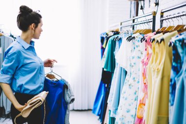 Profesyonel genç tasarımcı popüler giyim mağazasında çalışan yeni moda görüntüsü oluşturma. Seçme ve tarzı bir görünüm için elinde yetenekli kadın işçiye askıları trendy ile renkli tutan elbiseler