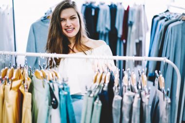 Kameraya bakarak ve raf arkasında trendy giyim duran Studio Tekstil giyim yeni koleksiyonu ile moda giysileri seçerken gülümseyen neşeli genç kadın müşteri yarım uzunluk portre