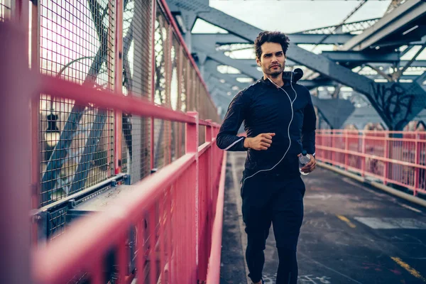 Professional male jogger dressed in active clothes running with modern earphones on bridge.Motivated fit man in sport clothes and headphones is engaged in sport workout and listening music