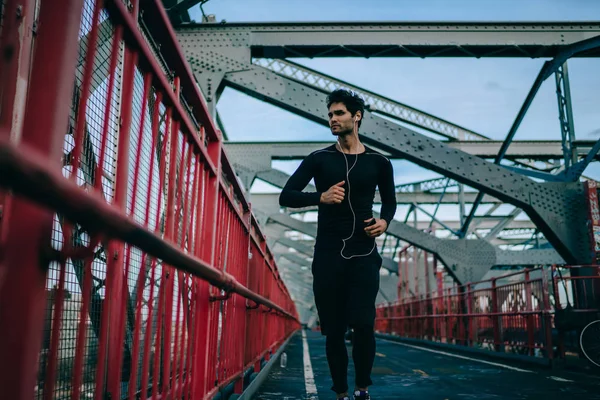 Motivated male jogger dressed in black active wear running and listening music player in earphones enjoying sport lifestyle.Sportsman in tracksuit training in urban setting on bridge