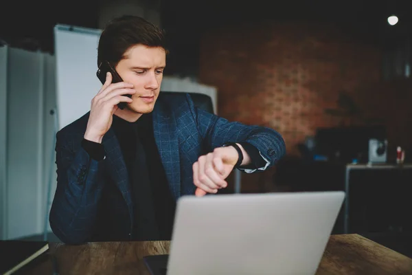 Serious male entrepreneur in formal wear looking at wristwatch during mobile phone conversation in cabinet, proud ceo arranging meeting through telephone talk checking time sitting near laptop