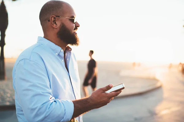 Bearded man sending text message via mobile telephone chatting online while enjoying summer evening, hipster guy satisfied with 4G connection in roaming share photos from vacation in social networks