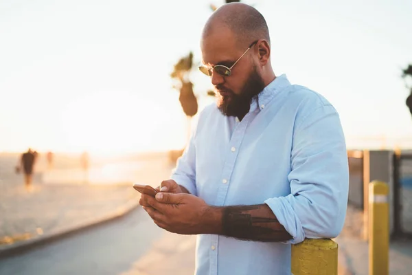 Handsome man in stylish outfit checking mail on smartphone app confirming booking on web page standing outdoors, bearded hipster guy blogging in social networks share files via modern mobile phone