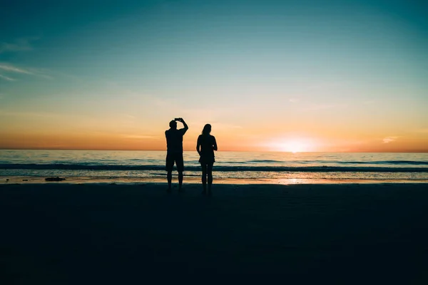 Male and female silhouettes on sandy shore of calm ocean during scenic sunset, couple in love enjoying beauty of evening tropical island view spending time on coast of sea during summer vacation