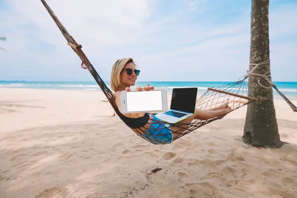 ビーチで夏の休暇の休日の間に現代のラップトップ コンピューターでリモートで作業中のアプリケーションまたは起業家の仕事の Web サイトに空白の画面を持つスマート フォンを示す女性観光客フリーランサー — ストック写真