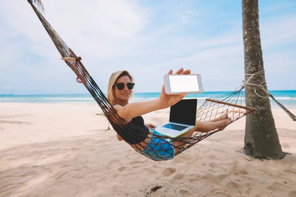 夏には熱帯の海海岸に休暇距離仕事中にハンモックにネットブックで働きながらインターネットの Web ページに空白の画面を持つスマート フォンを示す陽気な金髪女性フリーランサー — ストック写真