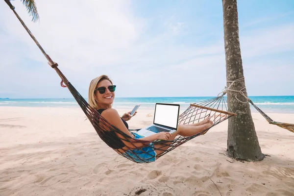現代のラップトップ コンピューターとハンモックに横たわってと熱帯の島の海岸での夏の休暇中にリモートで作業しながらカメラに微笑んでサングラスで幸せな若い女フリーランス起業家 — ストック写真