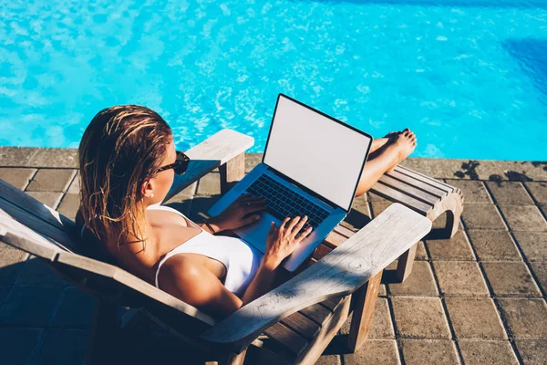 Female tourist freelancer typing information on keyboard of modern computer with blank copy space screen for your advertising information content working remotely and relaxing near pool at same time