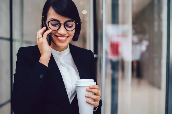 Positive female lawyer in black trendy suit discussing successful project during mobile conversation on smartphone device enjoying break with coffee to go outdoors nea publicity area