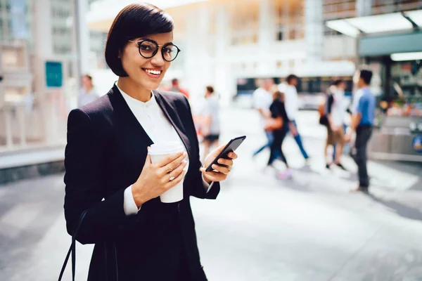 Portrait of successful female owner in elegant clothes and eyeglasses looking at camera chatting online on smartphone.Cheerful beautiful manager holding coffee to go standing near publicity area