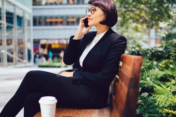 Female financial director of company in eyeglasses having phone conversation about work sitting on bench with coffee to go near office building.Manager in elegant wear talking on mobile during break