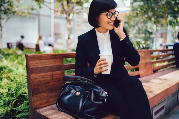 Successful female entrepreneur in eyewear discussing financial news on mobile phone while sitting on bench during work break with coffee in hands.Smiling businesswoman talking on cellular