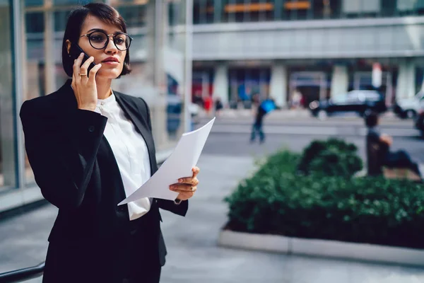 Confident female proud ceo in stylish elegant outfit solving financial problems during mobile conversation on telephone device standing outdoors near office building with paper accounting reports