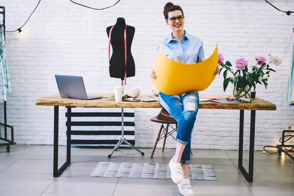 Happy professional seamstress in optical eyeglasses smiling at camera while holding yellow design sketch with mock up area in hands sitting on wooden desktop with modern computer and sewing tools