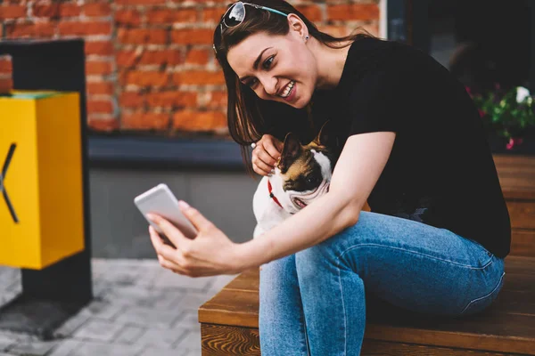 Cute positive female blogger trying to make funny selfie with little animal friend on smartphone camera for sharing in social networks.Smiling teenager taking photo of adorable puppy on telephone