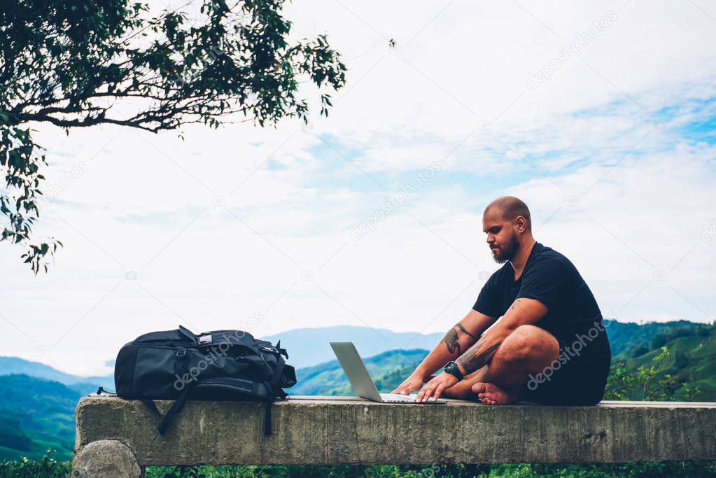Side view of pensive tourist working freelance at modern netbook doing distance job sitting outdoors in beautiful environment with high mountains on horizon during summer trip in faraway places