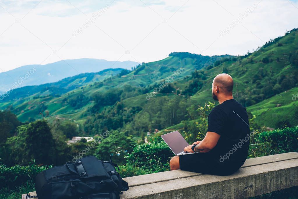 Young man freelancer sitting on hill with modern laptop computer for remote work while enjoying amazing view of high mountains with green plantation and vegetation during summer trip