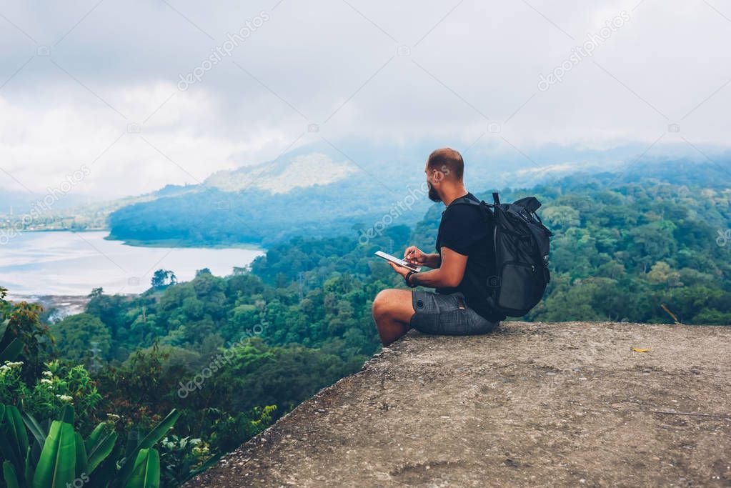 Skilled male painter climbed to top of mountain and depicts amazing scenery with green landscape of high hills and vegetation washed by sea sitting with backpack and using modern tablet for work