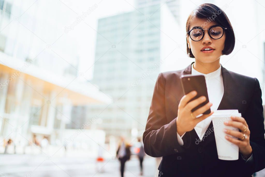Confident young entrepreneur in stylish spectacles browsing webpage for bank for checking account online on digital smartphone.Financial manager in formal wear and coffee in hand reading sms