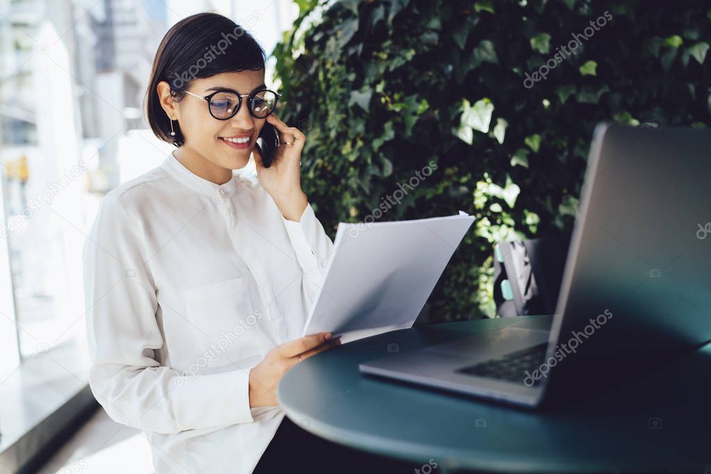 Cheerful owner of cafe in eyeglasses having phone conversation with colleague excited with good news.Successful manager satisfied with successful startup talking on mobile during checking project