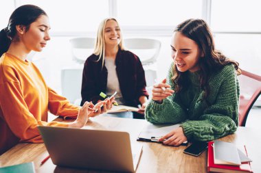 Birlikte ev ödevi geliştirmeye çalışan üç başarılı öğrenciler dizüstü aygıtı, şık coworking oturan proje. Kuruluş proje için öğretici izlerken gülmekten neşeli genç kadın
