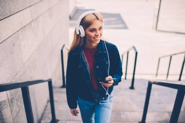 Kentsel ortamda geziniyor Kulaklığımda yabancı dil öğrenme ile dinleme ses ders sarı saçlı olan pozitif kadın. Neşeli öğrenci favori müzik keyfi denim ceket giymiş
