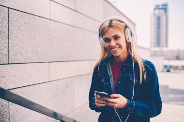 Yarım uzunluğu playlist ayakta kentsel ortamda gözde şarkı seçerken kameraya gülümseyen mutlu genç kadın portresi. Neşeli öğrenci dinleme ses kaseti yürüyüş sırasında modern Kulaklığımda