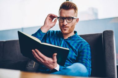 Skiled Kızıl saçlı genç adam portresi gözlük coworkin uzayda ilginç bir kitap okumak için sabit. Kahve dükkanında kapalı elinde ders kitabı ile oturan gözlük şık sahibi