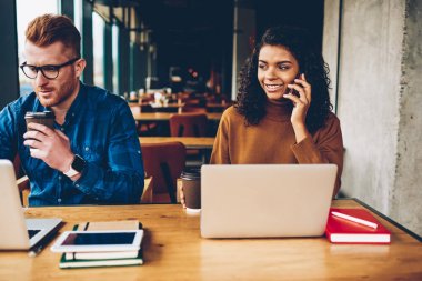 Smartphone ile birlikte dalgın Beyaz delikanlı kızıl saçlı ve kahve el ile 4 g dizüstü bilgisayar aygıt vasıl oturan ev ödevi üzerinde çalışırken konuşurken gülümseyen Afro-Amerikan öğrenci internet