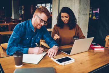 İki erkek ve kadın öğrencilerin ödev olarak defterini aşağı yazma ve modern dizüstü cihaz oturan eğitim semineri için hazırlanıyor bağlı kablosuz 4 g için kütüphane birlikte eğitim internet