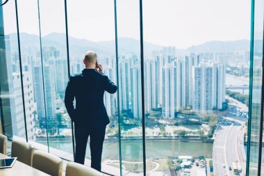 Ofiste ayakta ve metropolis harika manzarasını penceresinden bakarak ssuccessful iş projesi sonuçlarını smartphone aygıt yolu ile söz formal giyim erkek girişimci arkadan görünüm