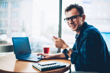 Half length portrait of handsome cheerful male developer in cool spectacles using modern technology during remote work in cafe.Smiling hipster guy looking at camera while updating software on laptop clipart