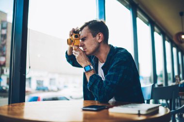 İyi düşünceli yetenekli fotoğrafçı rekreasyon vintage sarı kamera kapalı oturan resimleri yaparken zevk görünüyor. Serbest zaman sırasında fotoğraf çekmek smartwatch ile genç dalgın adam