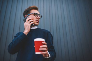 Yakışıklı dalgın hipster adam trendy Eyewear kentsel ayarında alan reklam ile duvar yanındaki yürüme. Promosyon arka plan karşı duran cep telefonunda konuşan genç iyi görünümlü adam