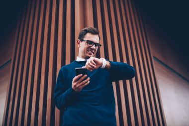 İyi görünümlü olumlu erkek öğrenci Street toplantı için beklenirken dijital smartwatch zamanında kontrol serin gözlükler. Zevk eğlence gülümseyen genç adam açık havada modern cihazlar ile