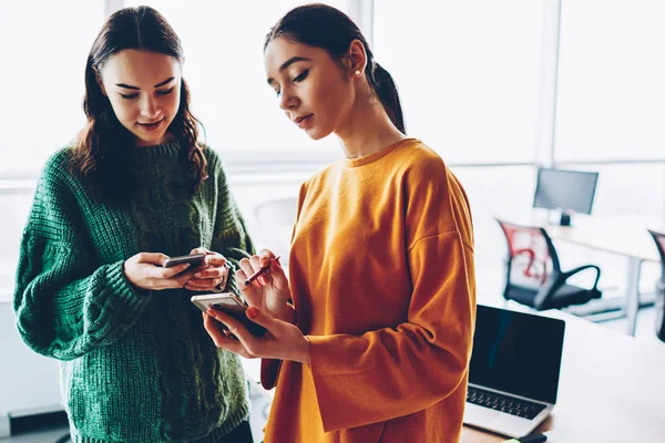 Friends discussing new posts in social networks on digital smartphones during break in office interior.Two female bloggers installing mobile apps on modern telephones standing indoors