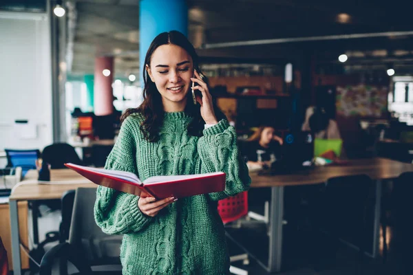 Positive brunette student talking with friends on smartphone while searching information in studying book.Successful young woman with textbook in hands laughing during mobile conversation on telephone