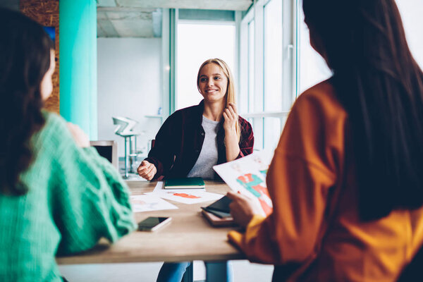 Successful young designer discussing plan of new sketches together with colleagues during briefing sitting at meeting desktop in office.Back view of young women collaborating on developing content