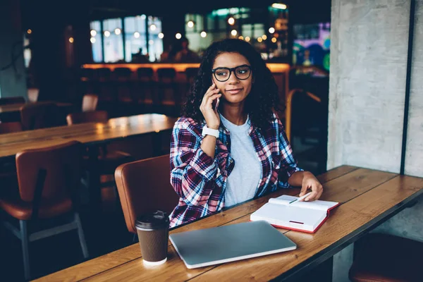 Afrika kökenli Amerikalı öğrenci şık gözlük ahşap masaüstü ile kahve ve netbook Üniversitesi'nde, Not Defteri ile oturma çalışma planı oluşturulurken smartphone cep telefonu operatörü ile konuşmak