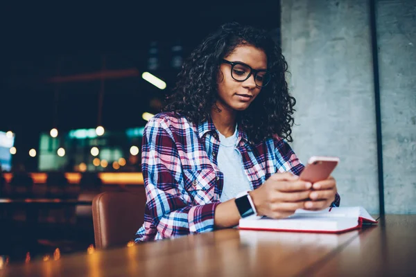 African american student messaging online on smartphone during studying in library.Dark skinned hipster girl in eyeglasses chatting with friend in social networks on cellular with 4G connection