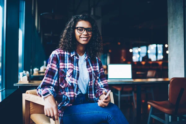 Muhteşem kıvırcık saçlı smartphone elinde tutarken kameraya gülümseyen başarılı afro Amerikan öğrenci portresi. Neşeli koyu tenli teen Kitaplığı'nda oturan şık gömlek giymiş