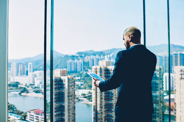Professional trader in formal wear checking bank account in an online database while talking with financial manager on smartphone standing in office near window with modern high skyscrapers outdoors