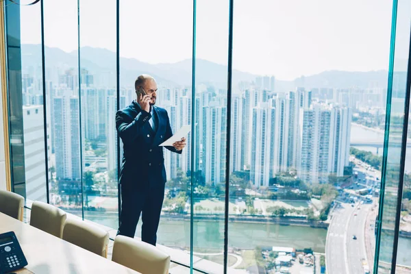 Smart financial director in black suit having serious mobile conversation with clients during checking accounting documentation in hands standing in office near window with metropolis view