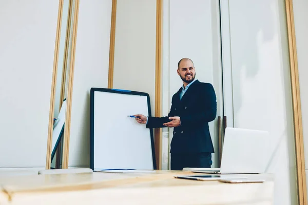 Successful male coach explaining training workshop standing near board with mock up area with your business information.Cheerful male trader in formal wear holds seminar session in coworking