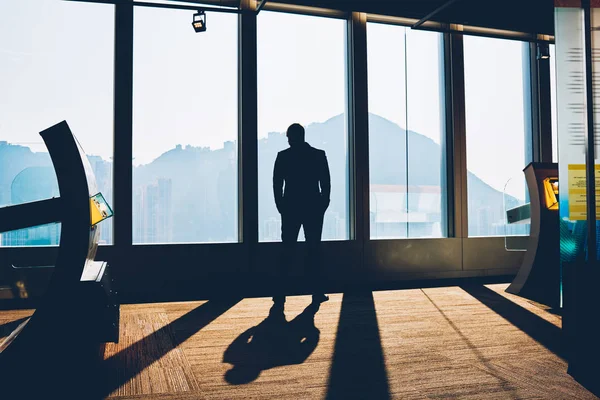 Back view of black silhouette of businessman dressed in formal wear standing in office of own financial company and looking out of window with beautiful scenery after hard day to thinking on new ideas