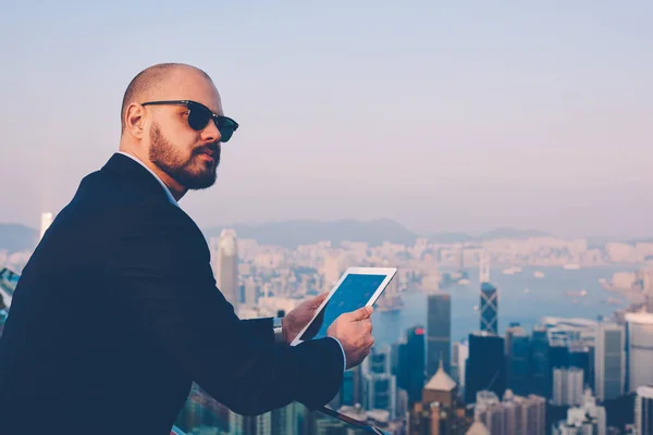 Side view of confident businessman in stylish sunglasses and elegant suit looking on side while updating media on digital touch pad enjoying metropolis view standing on balcony terrace