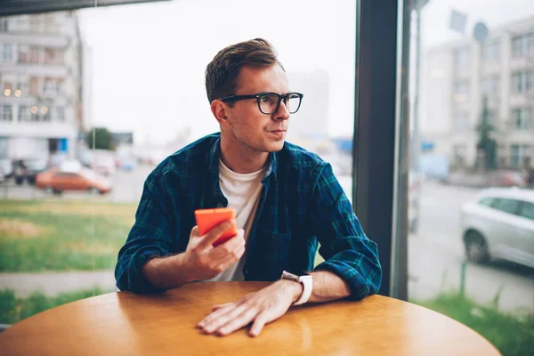 Young pondering creative male blogger in trendy eyeglasses looking away while waiting for meeting in coffee shop.Handsome hipster guy spending free time using modern technology sitting at table