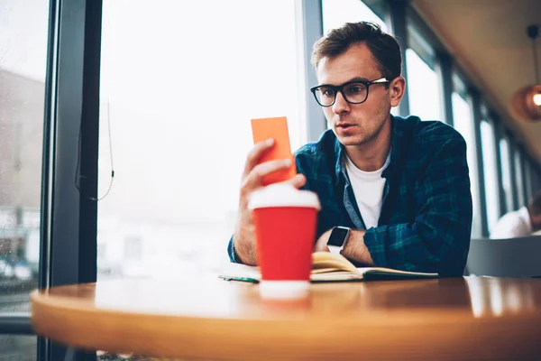 Young good looking male person in trendy eyeglasses surprised while searching information in browser via cellphone.Emotional hipster guy spending time indoors and watching live stream on website